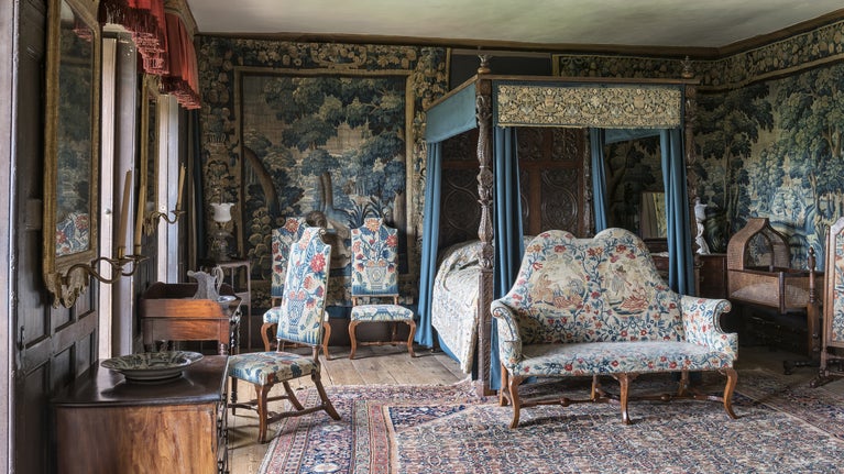 An elaborately decorated bedroom, with a four-poster bed, crib, three chairs, and a settee. The chairs and settee are upholstered in a decorative floral tapestry and there are tapestry wall hangings depicting trees.
